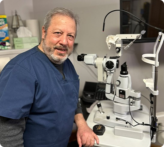 Dr. David Burstein, a Buffalo optometrist in blue scrubs, standing next to a slit lamp microscope in an exam room.