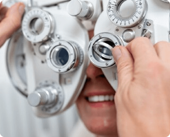A close-up shot of a patient behind a phoropter during an eye exam. An optician's hands are visible adjusting the dials and lenses of the equipment while the patient smiles.
