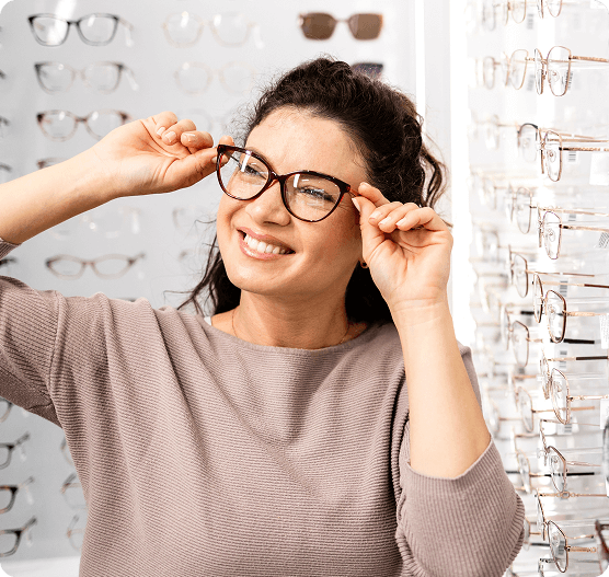A smiling woman trying on tortoiseshell eyeglasses in a bright optical shop with many frames on display.