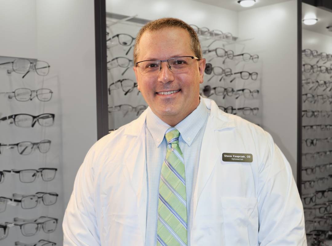 Dr. Steve Kasprzak, optometrist, smiling in a lab coat in front of an eyewear display at an eye care clinic.