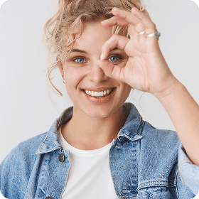 Smiling woman in a denim jacket making a circle with her fingers around one eye, suggesting clear or improved vision.