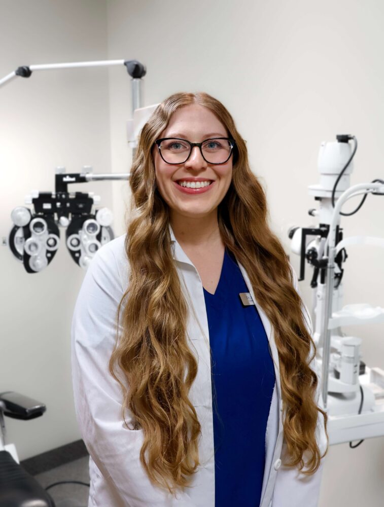 Dr. Jordan Lamb, Rochester NY optometrist, smiling in a white lab coat with eye exam equipment in the background.